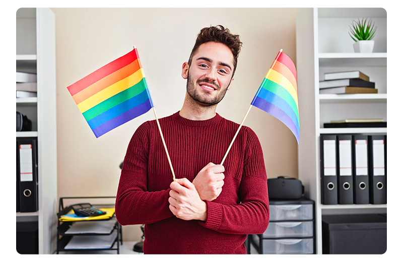 a guy wearing burgundy sweater holds 2 rainbow flags to celebrate Pride Month